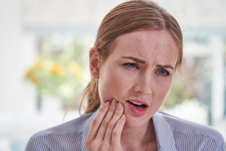 Woman with reddish hair tied back in a pony tail is holding her mouth in pain, likely experiencing a toothache.